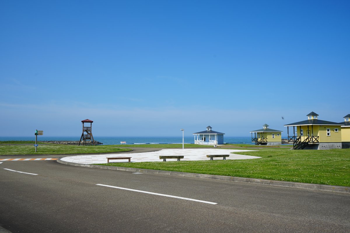 晴天の海沿い公園に並ぶカラフルなコテージと木製展望台、整備された芝生と道路