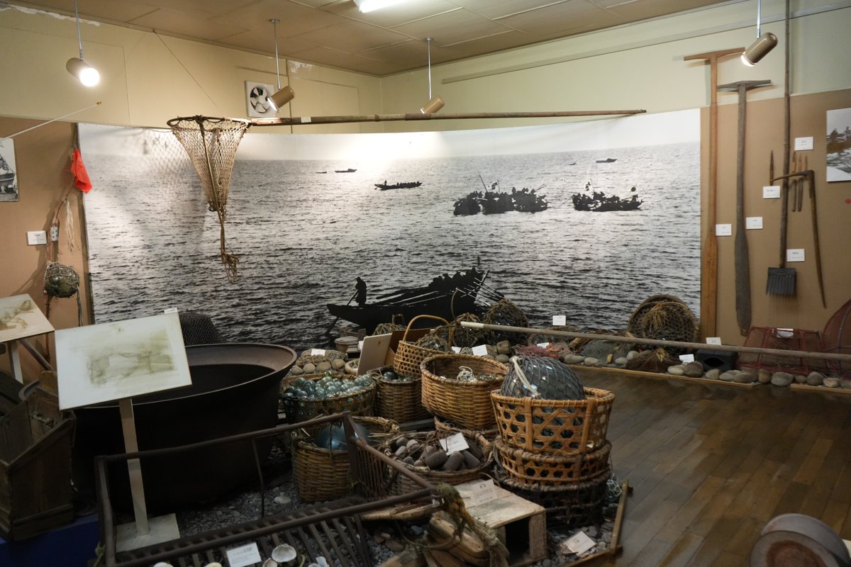 漁業博物館の展示室。かつての漁船の白黒写真パネルの前に、籠・漁網・浮き球など多数の漁具が並ぶ展示の様子