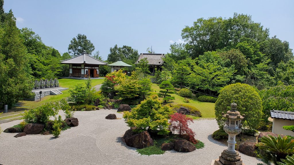 石灯籠と刈り込まれた植木が並ぶ枯山水庭園、奥に寺院の建物が見える風景