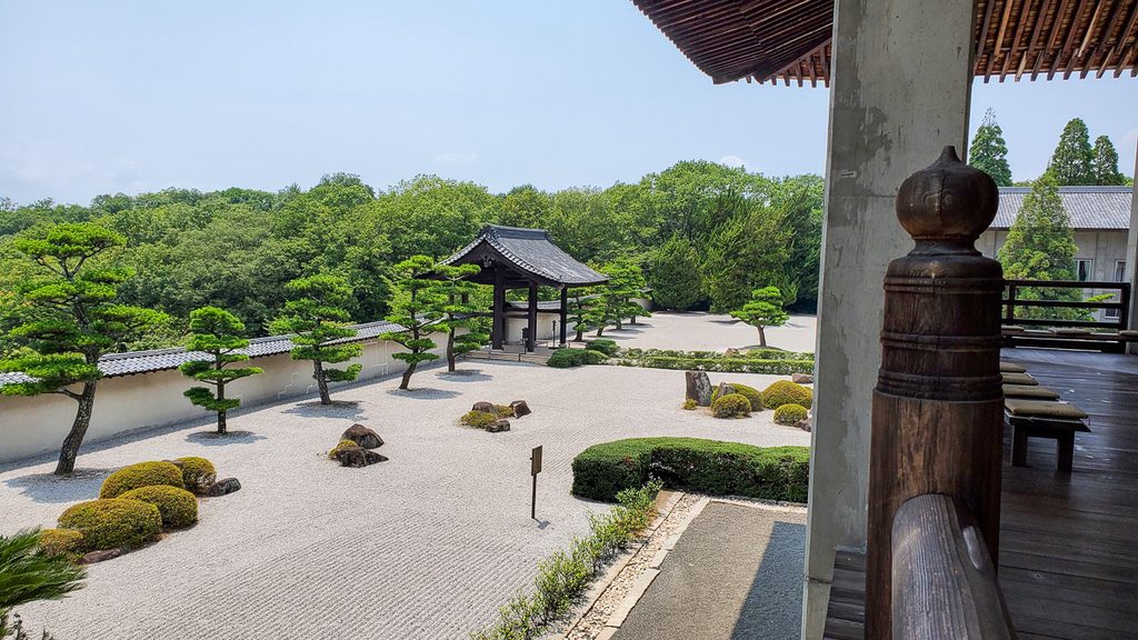 白砂に松と石を配した枯山水の庭と、奥に佇む木造の門、周囲を囲む緑の木々。